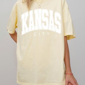Peut inclure: Un t-shirt &agrave; manches courtes jaune p&acirc;le avec le texte KANSAS CITY imprim&eacute; en grandes lettres blanches sur la poitrine. Le v&ecirc;tement pr&eacute;sente une encolure ronde et une coupe d&eacute;contract&eacute;e, avec le mot KANSAS arqu&eacute; au-dessus du mot CITY.