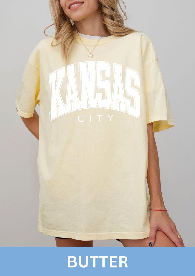 Peut inclure: Un t-shirt &agrave; manches courtes jaune p&acirc;le avec le texte KANSAS CITY imprim&eacute; en grandes lettres blanches sur la poitrine. Le v&ecirc;tement pr&eacute;sente une encolure ronde et une coupe d&eacute;contract&eacute;e, avec le mot KANSAS arqu&eacute; au-dessus du mot CITY.