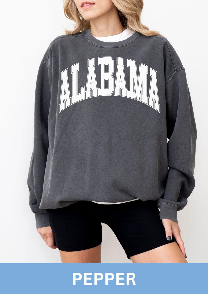 May include: A charcoal grey crewneck sweatshirt featuring the word ALABAMA printed in large white block letters across the chest in an arched design. The garment has long sleeves with ribbed cuffs and a ribbed neckline, worn over a white shirt.