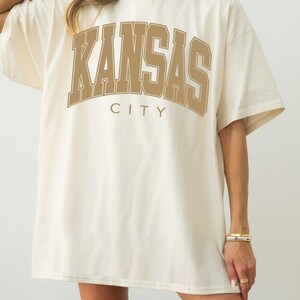 Peut inclure: Un t-shirt &agrave; manches courtes ivoire avec le texte KANSAS CITY imprim&eacute; en grandes lettres arqu&eacute;es de couleur beige dans un style universitaire sur la poitrine. Le v&ecirc;tement pr&eacute;sente un col rond et une coupe ample avec des &eacute;paules tombantes.