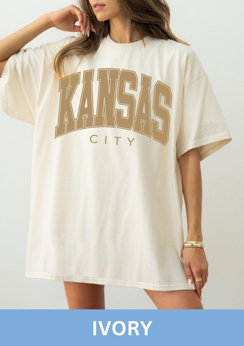 Peut inclure: Un t-shirt &agrave; manches courtes ivoire avec le texte KANSAS CITY imprim&eacute; en grandes lettres arqu&eacute;es de couleur beige dans un style universitaire sur la poitrine. Le v&ecirc;tement pr&eacute;sente un col rond et une coupe ample avec des &eacute;paules tombantes.
