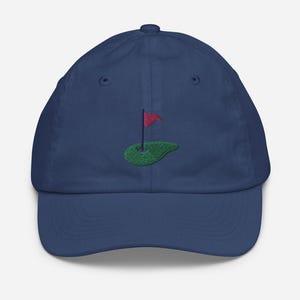 May include: A navy blue baseball cap with an embroidered green golf course and a red flag.