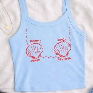 Puede incluir: Camiseta sin mangas azul claro con gr&aacute;ficos de conchas rojas y texto. El texto dice "MARY'S BACH MIAMI JULY 2026". La camiseta tiene tirantes finos y cuello redondo. La tela parece suave.