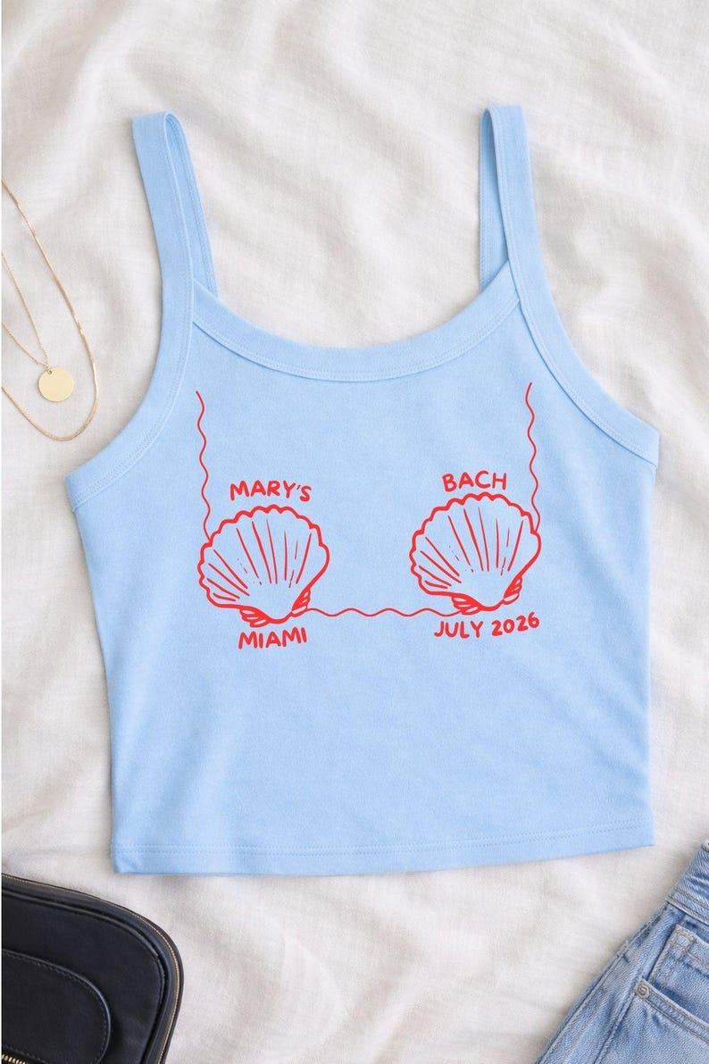 Puede incluir: Camiseta sin mangas azul claro con gr&aacute;ficos de conchas rojas y texto. El texto dice "MARY'S BACH MIAMI JULY 2026". La camiseta tiene tirantes finos y cuello redondo. La tela parece suave.