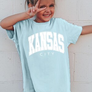 Peut inclure: Un t-shirt &agrave; manches courtes bleu clair avec le texte KANSAS CITY imprim&eacute; en grandes lettres blanches de style universitaire sur la poitrine. Le v&ecirc;tement pr&eacute;sente un col rond et une coupe d&eacute;contract&eacute;e en tissu de couleur chambray.