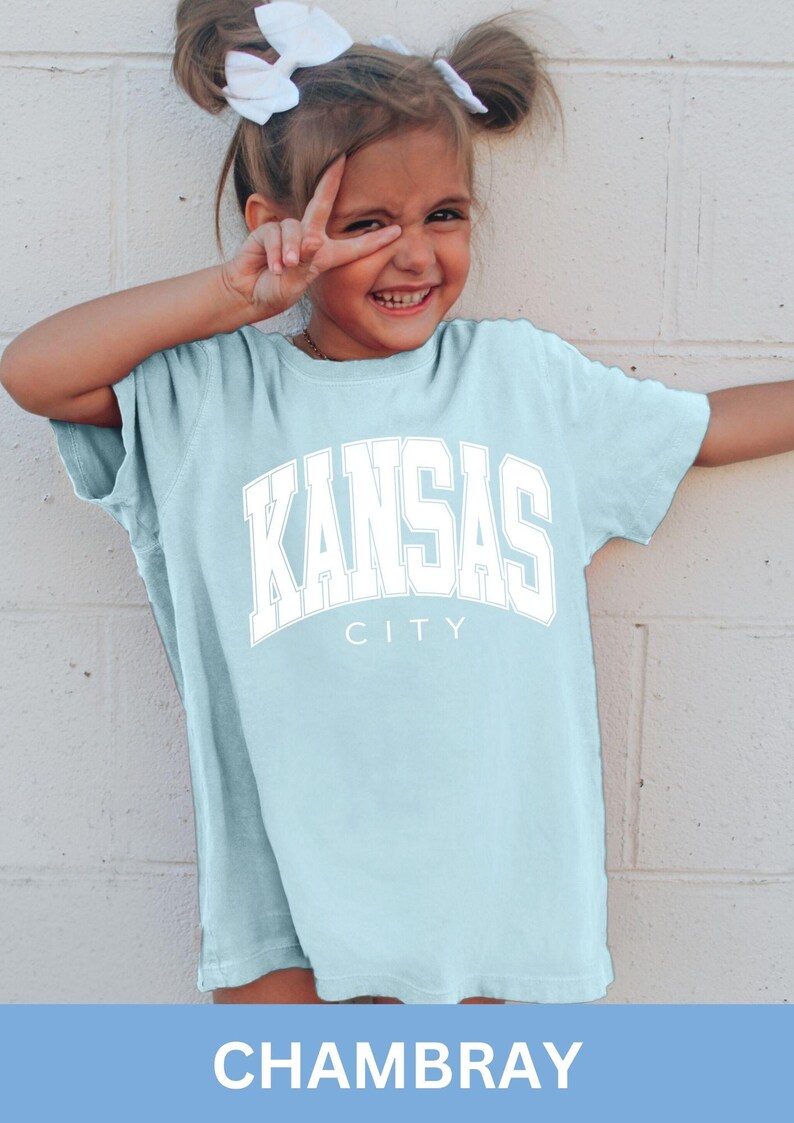 Peut inclure: Un t-shirt &agrave; manches courtes bleu clair avec le texte KANSAS CITY imprim&eacute; en grandes lettres blanches de style universitaire sur la poitrine. Le v&ecirc;tement pr&eacute;sente un col rond et une coupe d&eacute;contract&eacute;e en tissu de couleur chambray.