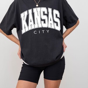 Peut inclure: Un t-shirt noir &agrave; manches courtes avec le texte blanc KANSAS CITY dans une police universitaire audacieuse sur la poitrine, superpos&eacute; sur un sous-v&ecirc;tement blanc, associ&eacute; &agrave; un short de cycliste noir c&ocirc;tel&eacute; et un collier &agrave; cha&icirc;ne dor&eacute;e.