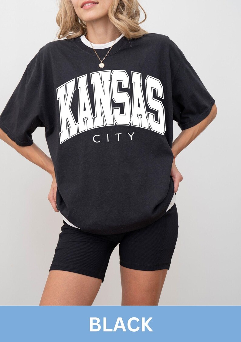 Peut inclure: Un t-shirt noir &agrave; manches courtes avec le texte blanc KANSAS CITY dans une police universitaire audacieuse sur la poitrine, superpos&eacute; sur un sous-v&ecirc;tement blanc, associ&eacute; &agrave; un short de cycliste noir c&ocirc;tel&eacute; et un collier &agrave; cha&icirc;ne dor&eacute;e.