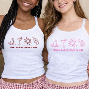 May include: Two women wearing white tank tops with a pink graphic and text that reads "MARY'S BACH/ MIAMI, FL 2026". They are also wearing red and white checkered pajama shorts. The tank tops have a bikini, cocktail, sun, and seashell design.