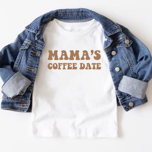May include: White t-shirt with brown text that reads "Mama's Coffee Date" in a retro font. The shirt is folded and displayed on top of a blue denim jacket.