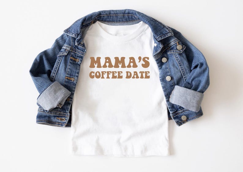 May include: White t-shirt with brown text that reads "Mama's Coffee Date" in a retro font. The shirt is folded and displayed on top of a blue denim jacket.
