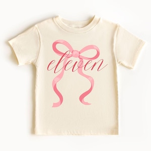 May include: Cream-colored t-shirt featuring a pink bow design with the word "eleven" in a cursive font. The bow has flowing ribbons. The shirt is made of a soft material.