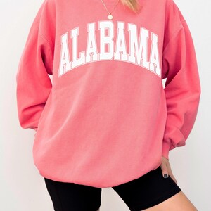 May include: A watermelon pink crewneck sweatshirt featuring the word ALABAMA printed in large white capital letters across the chest in an arched collegiate font. The garment has long sleeves and a ribbed neckline, worn over a white undershirt.