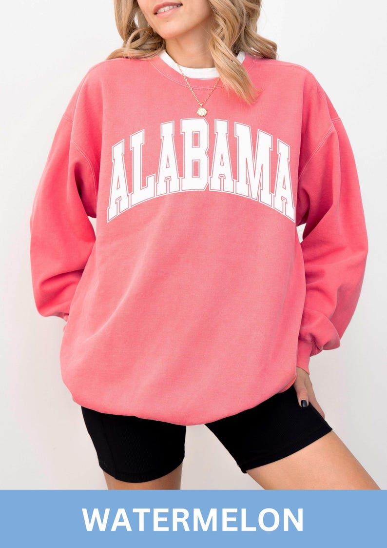 May include: A watermelon pink crewneck sweatshirt featuring the word ALABAMA printed in large white capital letters across the chest in an arched collegiate font. The garment has long sleeves and a ribbed neckline, worn over a white undershirt.