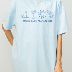May include: Light blue t-shirt with a blue graphic of a bikini, cocktail, sun, and seashell. The text "MARY'S BACH/ MIAMI, FL 2026" is printed below the graphics. The shirt is a casual, oversized fit.