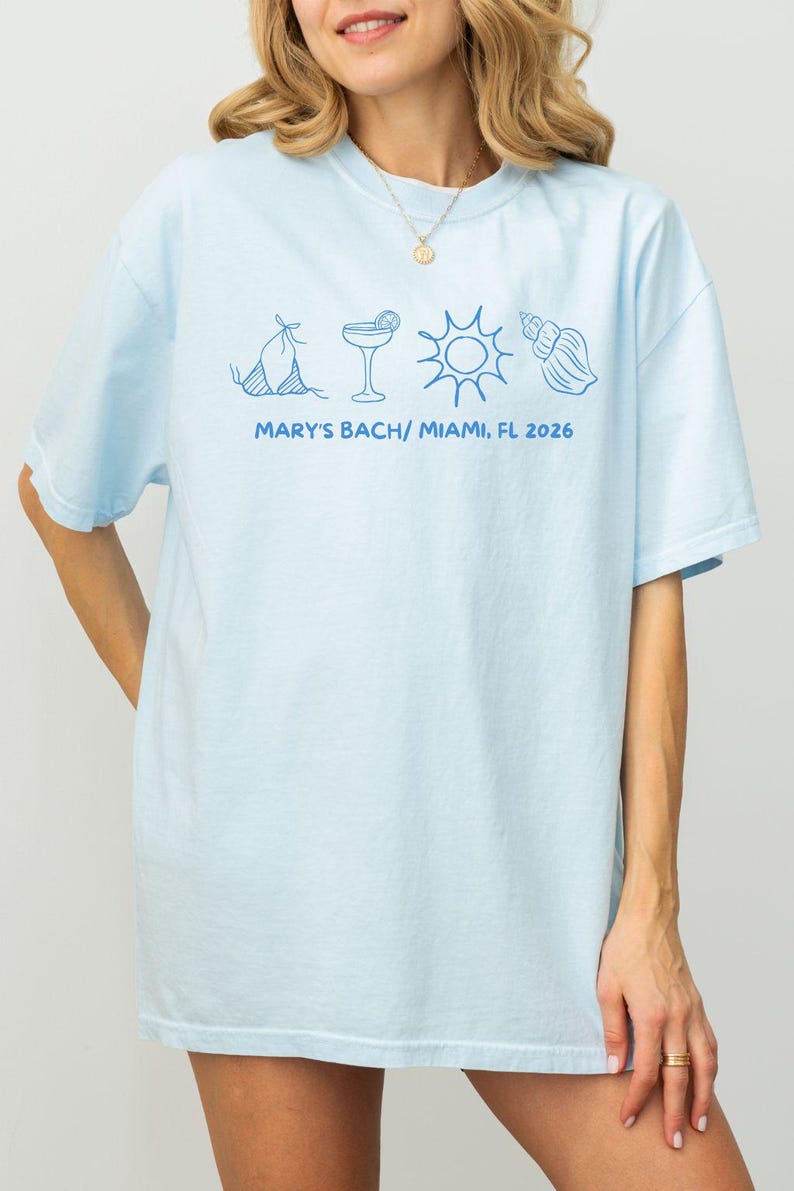 May include: Light blue t-shirt with a blue graphic of a bikini, cocktail, sun, and seashell. The text "MARY'S BACH/ MIAMI, FL 2026" is printed below the graphics. The shirt is a casual, oversized fit.