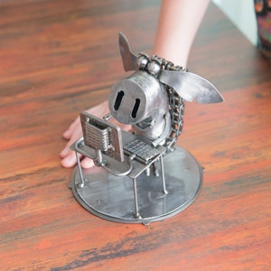 Könnte beinhalten: Eine Metallskulptur eines Schweins, das an einem Schreibtisch mit einem Computer und einer Tastatur sitzt. Das Schwein ist aus recyceltem Metall gefertigt und hat einen skurrilen, cartoonartigen Stil.