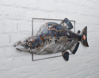 Puede incluir: Escultura metálica de un pez, montada en una pared de ladrillos blancos. El pez está construido con varias piezas de metal, con una combinación de colores azul y plata. La escultura está enmarcada por un cuadrado de metal plateado.