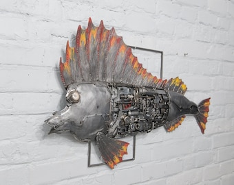 Puede incluir: Una escultura de pez de metal con una aleta roja, naranja y amarilla. El pez está hecho de varias piezas de metal, incluyendo engranajes y tornillos. El pez está montado en una pared de ladrillo blanco.
