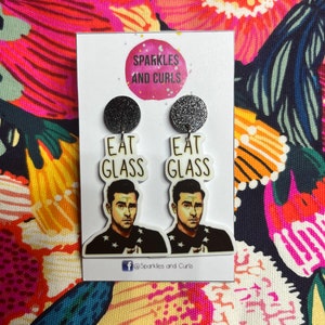 May include: Novelty earrings with black glitter tops and white rectangular bottoms. The earrings feature a portrait of a man and the words "EAT GLASS". The earrings are attached to a white card with the text "Sparkles and Curls".