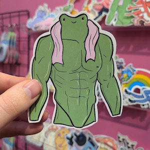 May include: A green frog sticker with a muscular physique and a pink towel. The frog has a smiling face and a cartoon-like design. Other stickers are visible in the background, with a pink wall behind them.
