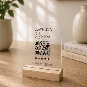 May include: A clear rectangular acrylic sign with a wooden base. The sign features black text that reads LEAVE US A, Review, a QR code, five black stars, and the text YOUR BUSINESS NAME. The sign is upright and displays a professional design.
