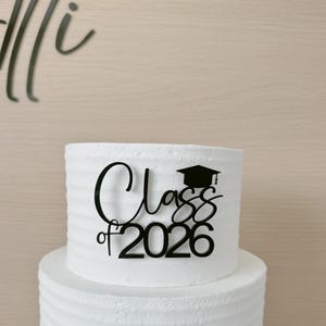 May include: A two-tiered white cake with textured frosting. A black topper reads "Class of 2026" with a graduation cap icon. The cake is set against a light wood-grain background, suggesting a celebratory occasion.