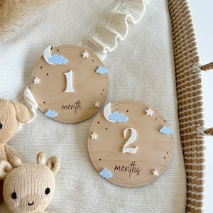 May include: Two wooden baby milestone discs featuring a moon, stars, and clouds design. The first disc reads "1 month" and the second disc reads "2 months".