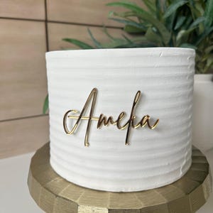 May include: White cake with gold lettering that says "Amelia" on top of a gold cake stand.