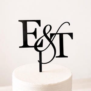 May include: A black acrylic cake topper featuring the letters E and T in a bold serif font, connected by a stylized ampersand symbol in the center. The topper is inserted into the top of a plain white frosted cake with a smooth texture.