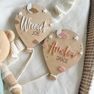 Pode incluir: Duas placas de nome de madeira em forma de bal&atilde;o de ar quente. Uma apresenta os nomes "Wrenick" e "Joe" em letras brancas, com estrelas brancas, uma lua e nuvens castanhas. A outra diz "Amelia" e "Grace" em rosa, com decora&ccedil;&otilde;es semelhantes.