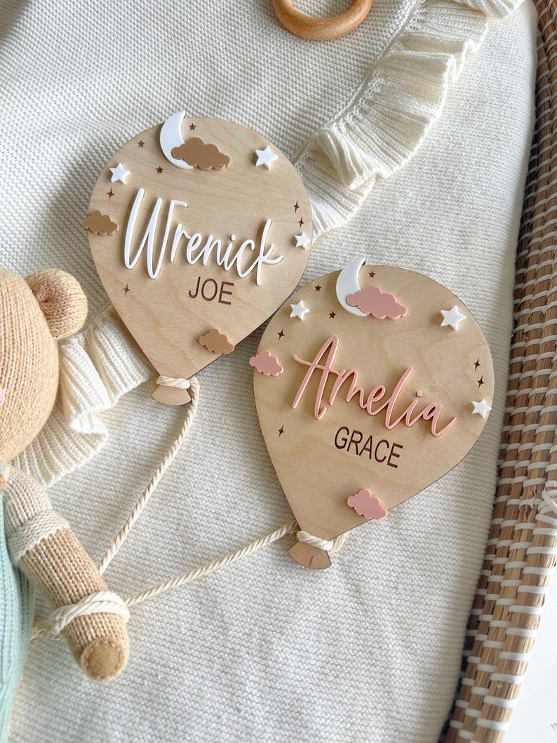 Pode incluir: Duas placas de nome de madeira em forma de bal&atilde;o de ar quente. Uma apresenta os nomes "Wrenick" e "Joe" em letras brancas, com estrelas brancas, uma lua e nuvens castanhas. A outra diz "Amelia" e "Grace" em rosa, com decora&ccedil;&otilde;es semelhantes.