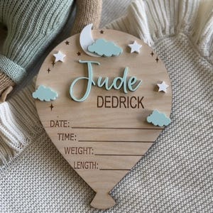 Pode incluir: Placa de madeira em forma de bal&atilde;o com o nome "Jude Dedrick" em fonte cursiva. A placa apresenta uma lua, nuvens e estrelas. Inclui espa&ccedil;os para registrar data, hora, peso e comprimento.