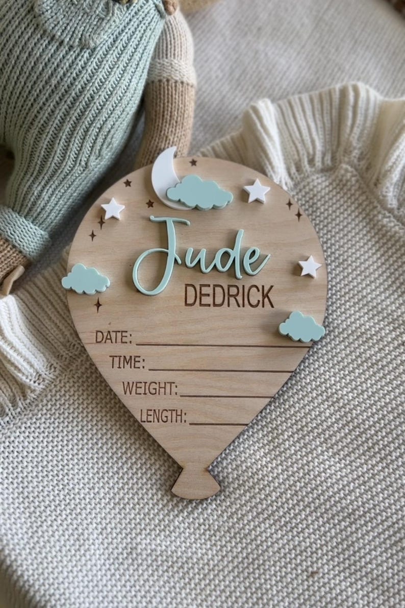 Pode incluir: Placa de madeira em forma de bal&atilde;o com o nome "Jude Dedrick" em fonte cursiva. A placa apresenta uma lua, nuvens e estrelas. Inclui espa&ccedil;os para registrar data, hora, peso e comprimento.