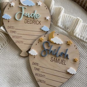 Pode incluir: Duas placas de madeira em forma de bal&atilde;o para anunciar o nascimento de um beb&eacute;. Uma diz "Jude Dedrick" com uma lua, nuvens e estrelas. A outra diz "Shiloh Samuel" com uma lua, nuvens e estrelas. Cada uma tem linhas para data, hora, peso e comprimento.