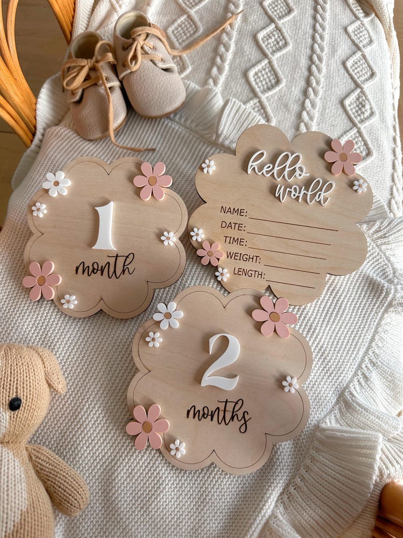 May include: Two wooden baby milestone signs decorated with pink and white flowers. The first sign reads "1 month" and the second sign reads "2 months". A third sign reads "hello world" with spaces for baby's name, date, time, weight, and length.