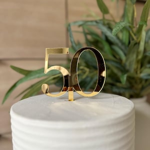 May include: Gold-colored number 50 cake topper. The number is made of a reflective material. The topper is on top of a white cake with a textured frosting.