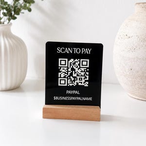 May include: A black rectangular payment sign with white text that reads SCAN TO PAY, PAYPAL, and $BUSINESSPAYPALNAME, featuring a central white QR code. The sign is held upright by a rectangular light-colored wooden base stand.