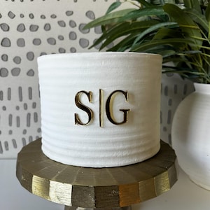 May include: A white, cylindrical ceramic piece with the gold letters "S|G" on the front, resting on a gold-colored, geometric pedestal. The piece has a textured, ridged surface. Decorative greenery is visible in the background.