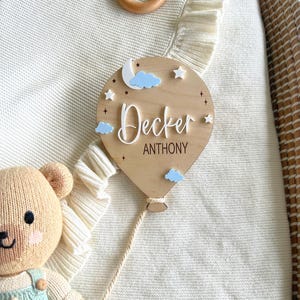 Pode incluir: Uma placa de nome em forma de bal&atilde;o de ar quente de madeira com o nome "Decker Anthony" em escrita branca. A placa apresenta nuvens azuis, estrelas e uma lua crescente. Um ursinho de pel&uacute;cia bege usando macac&atilde;o azul est&aacute; no canto inferior esquerdo.