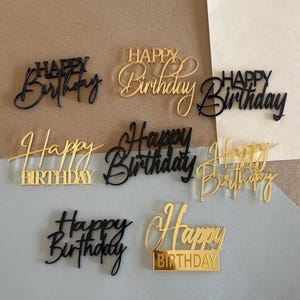 May include: A collection of gold and black acrylic cake toppers with the words "Happy Birthday" in various elegant fonts. The toppers are in different styles, some with a script font and others with a more blocky design.