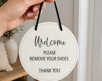 May include: A circular off-white hanging sign with a thin black border and black text that reads Welcome, PLEASE REMOVE YOUR SHOES, THANK YOU. The sign is suspended by a thin black cord attached to the top, featuring a clean and simple design.
