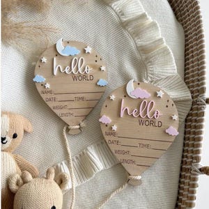 Pode incluir: Duas placas de an&uacute;ncio de nascimento em forma de bal&atilde;o de madeira com as palavras "hello world". Cada placa tem espa&ccedil;o para nome, data, hora, peso e comprimento. Nuvens decorativas, estrelas e uma lua acentuam o design. Um urso de pel&uacute;cia de malha est&aacute; no fundo.