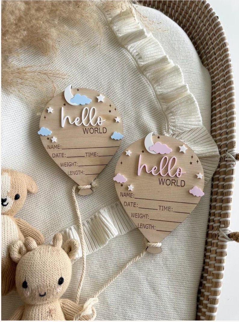 Pode incluir: Duas placas de an&uacute;ncio de nascimento em forma de bal&atilde;o de madeira com as palavras "hello world". Cada placa tem espa&ccedil;o para nome, data, hora, peso e comprimento. Nuvens decorativas, estrelas e uma lua acentuam o design. Um urso de pel&uacute;cia de malha est&aacute; no fundo.