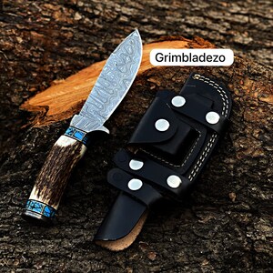Może przedstawiać: N&oacute;ż myśliwski z wzorzystą stalową klingą i brązową rękojeścią z poroża jelenia z turkusowymi akcentami. N&oacute;ż znajduje się w czarnej sk&oacute;rzanej pochwie z zatrzaskiem i szwami. Tekst "Grimbladezo" jest widoczny na zdjęciu.