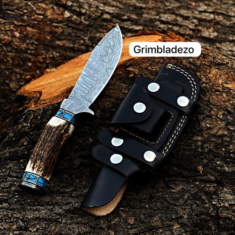 Może przedstawiać: N&oacute;ż myśliwski z wzorzystą stalową klingą i brązową rękojeścią z poroża jelenia z turkusowymi akcentami. N&oacute;ż znajduje się w czarnej sk&oacute;rzanej pochwie z zatrzaskiem i szwami. Tekst "Grimbladezo" jest widoczny na zdjęciu.