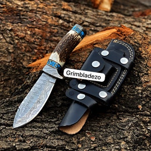 Może przedstawiać: N&oacute;ż myśliwski ze stali damasceńskiej z rękojeścią z poroża jelenia i akcentami turkusowymi. N&oacute;ż znajduje się w czarnej sk&oacute;rzanej pochwie z zatrzaskiem i napisem "Grimbladezo" z przodu.