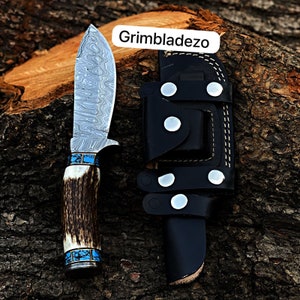 Może przedstawiać: N&oacute;ż myśliwski z wzorzystą stalową klingą, brązową rękojeścią z poroża z turkusowymi akcentami i czarną sk&oacute;rzaną pochwą z srebrnymi nitami. Pochwa ma napis "Grimbladezo".