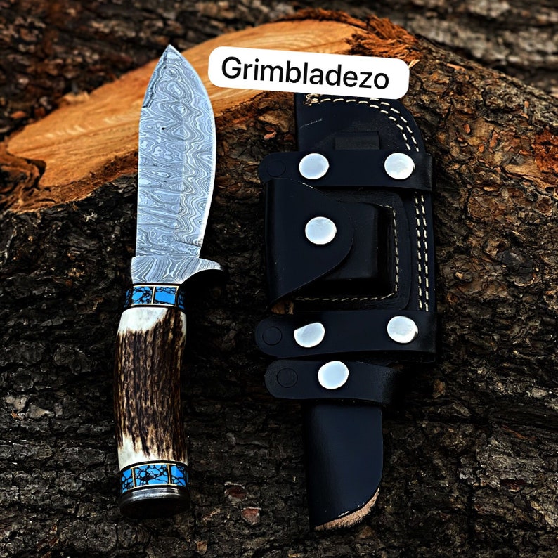 Może przedstawiać: N&oacute;ż myśliwski z wzorzystą stalową klingą, brązową rękojeścią z poroża z turkusowymi akcentami i czarną sk&oacute;rzaną pochwą z srebrnymi nitami. Pochwa ma napis "Grimbladezo".