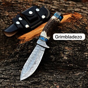 Może przedstawiać: N&oacute;ż myśliwski z ostrzem ze stali damasceńskiej i rękojeścią z poroża jelenia. Rękojeść zdobiona jest turkusowymi intarsjami. N&oacute;ż leży na kawałku drewna. Grimbladezo.